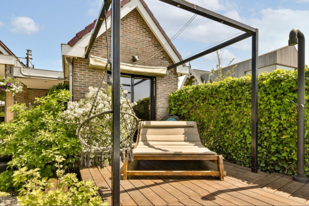 A long wooden deck with a lawn chair on top and green plants surrounding. 