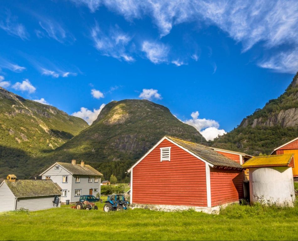 A red barn on a big meadow with other structures and mountains a distance away.